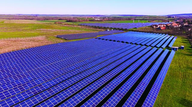 Wide aerial view of solar farm near a village. Rows of blue panels stretch toward rooftops and open countryside under clear sky.