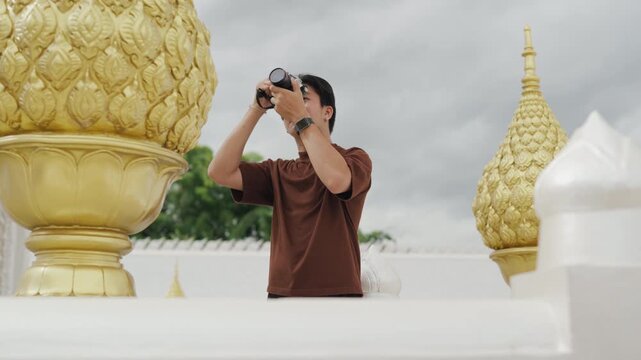 Handsome young Asian male tourist taking beautiful photos with digital camera at white temple. Cheerful solo traveler enjoying cultural sightseeing adventure on summer vacation.