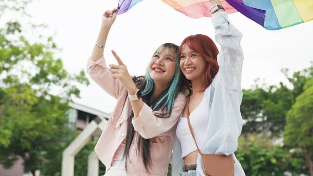 Beautiful Asian lesbian couple holding a rainbow flag and embracing in green park. Cheerful young women celebrating LGBTQ pride month equality and expressing romantic love.