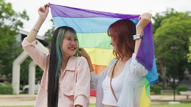 Beautiful Asian lesbian couple holding a rainbow flag and embracing in green park. Cheerful young women celebrating LGBTQ pride month equality and expressing romantic love.