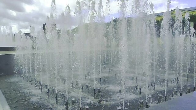 Water fountains splashing in a city park under a cloudy sky, urban landscape, refreshing summer day, public water feature, outdoor scenery.