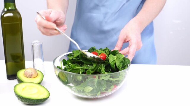 Hands toss salad greens in a clear bowl. Fresh spinach and cherry tomatoes mix with dressing. Avocado halves sit beside olive oil. Person wears blue apron, focused on preparation