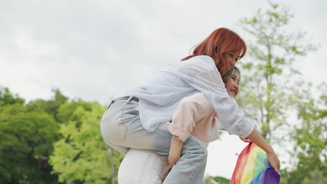 Beautiful Asian lesbian couple holding a rainbow flag and embracing in green park. Cheerful young women celebrating LGBTQ pride month equality and expressing romantic love.