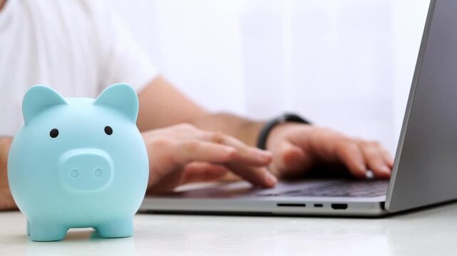 Light blue piggy bank sits near laptop. Hands type on keyboard, focused on work. Soft lighting suggests calm, productive environment. Ideal for finance, budgeting, or savings concepts