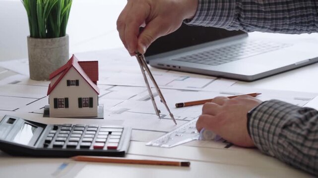Hand draws plans with compass near miniature home. Calculator and laptop sit beside the workspace. Desk holds blueprint and small house model. Professional mood, focused on design and budgeting