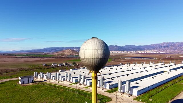 Spherical water tower overlooking poultry farm. Yellow pole and round tank rise above rows of barns with mountains behind.