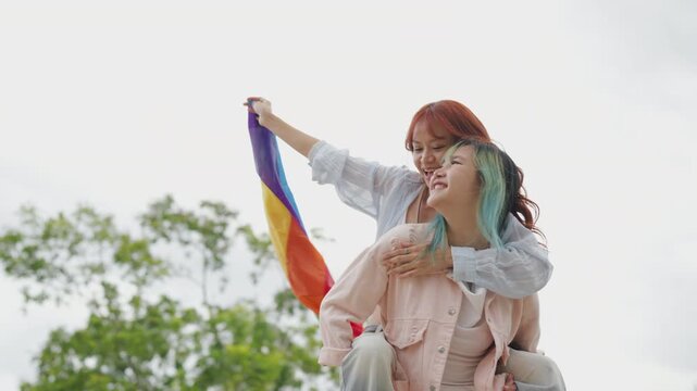 Beautiful Asian lesbian couple holding a rainbow flag and embracing in green park. Cheerful young women celebrating LGBTQ pride month equality and expressing romantic love.