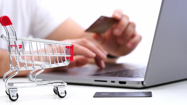 Hands hold card near laptop. Mini cart sits beside screen. User shops online, paying with card. Ideal for e-commerce, finance, retail scenes. No animals or plants in image