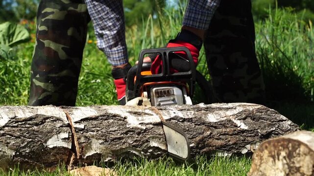 Worker cuts log with chainsaw on grassy field. Person wears camouflage pants and red gloves. Chainsaw rests on cut bark, ready for next slice. Sunlight highlights green grass and wood texture