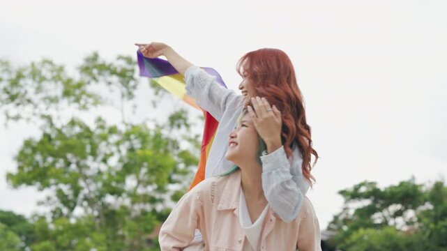 Beautiful Asian lesbian couple holding a rainbow flag and embracing in green park. Cheerful young women celebrating LGBTQ pride month equality and expressing romantic love.
