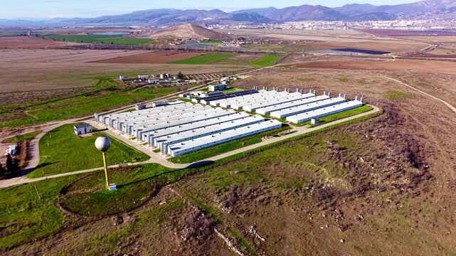 Wide aerial view of poultry farm complex with water tower. Rows of barns and silos sit beside a road with distant mountains.