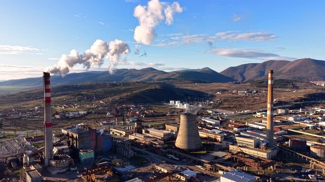 Industrial cooling tower and chimney with steam. White smoke drifts above a factory site framed by mountains and rooftops.