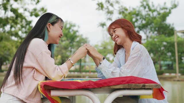 Beautiful Asian lesbian couple holding a rainbow flag and embracing in green park. Cheerful young women celebrating LGBTQ pride month equality and expressing romantic love.