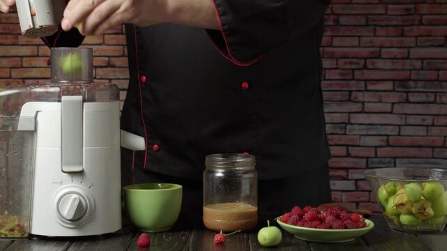 Male chef in black apron adjusts juicer settings while preparing fresh fruit ingredients, including strawberries and green apples, on a wooden table against a rustic brick wall backdrop