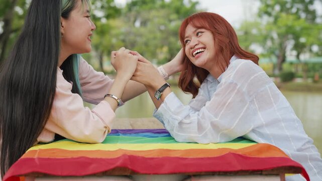 Beautiful Asian lesbian couple holding a rainbow flag and embracing in green park. Cheerful young women celebrating LGBTQ pride month equality and expressing romantic love.