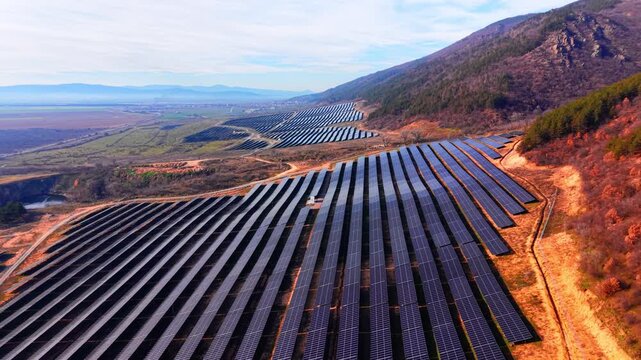 Aerial view of solar array descending mountain slope. Panel rows extend toward a village and farmland valley beneath forested peaks.