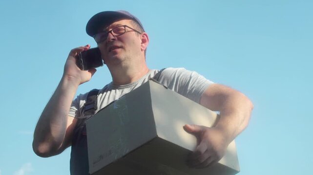 Postman in gray shirt and blue cap holds a cardboard box while talking on the phone outdoors, transitioning to checking his smartphone, with a clear blue sky in the background