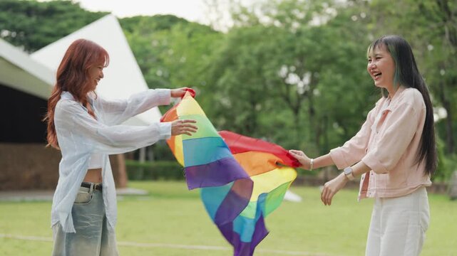 Happy young Asian lesbian couple sitting on a rainbow flag enjoying romantic summer picnic in park. Beautiful LGBTQ women sharing lovely weekend date and relaxing lifestyle.