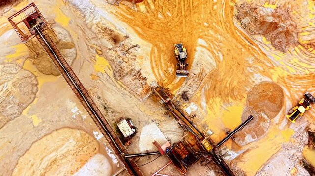 Aerial view of loader and conveyor at sand pit. Overhead angle shows a wheel loader beside rusted conveyor structures and sand mounds.