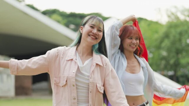 Happy young Asian lesbian couple sitting on a rainbow flag enjoying romantic summer picnic in park. Beautiful LGBTQ women sharing lovely weekend date and relaxing lifestyle.