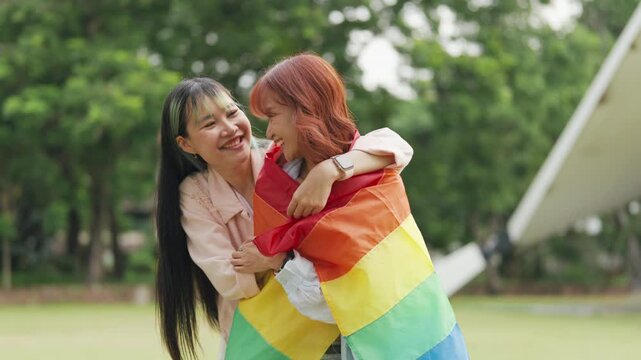Happy young Asian lesbian couple sitting on a rainbow flag enjoying romantic summer picnic in park. Beautiful LGBTQ women sharing lovely weekend date and relaxing lifestyle.