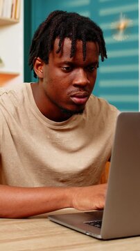 African American young man on home table typing on laptop with focused face during remote work at home. Black boy on home table doing online job planning project using computer in living room interior