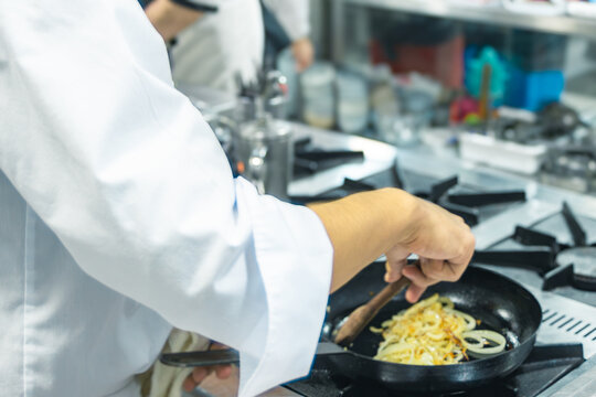 Chef stirring caramelized onions in a frying pan on a professional gas stove, focusing on healthy global cuisine and innovative gastronomy for sustainable practices