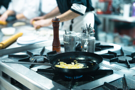 Chefs are actively preparing food in a professional kitchen, with sliced onions sauteing in a pan on a gas stove, symbolizing healthy and creative gastronomy