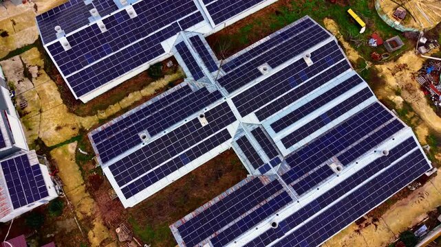 Drone shot of solar farm rooftop grid, rural Bulgaria. Overhead angle showing dark photovoltaic modules, rusty siding and overgrown yard between structures.