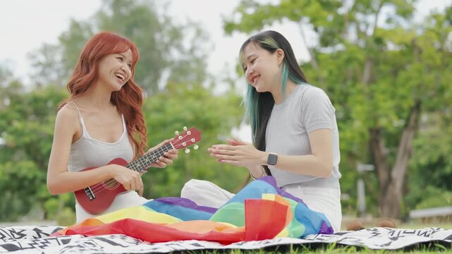 Cheerful Asian lesbian couple playing acoustic guitar and singing together in a sunny green park. Happy young LGBTQ women enjoying romantic musical date and summer freedom.