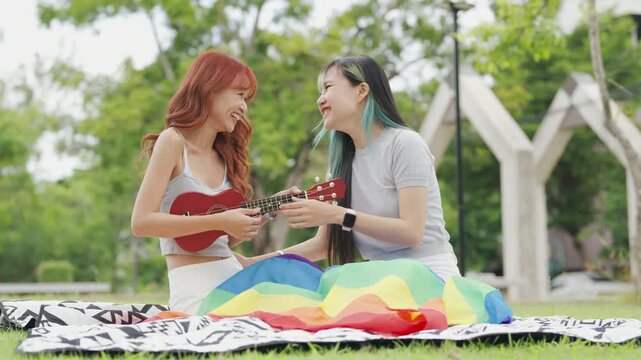 Cheerful Asian lesbian couple playing acoustic guitar and singing together in a sunny green park. Happy young LGBTQ women enjoying romantic musical date and summer freedom.