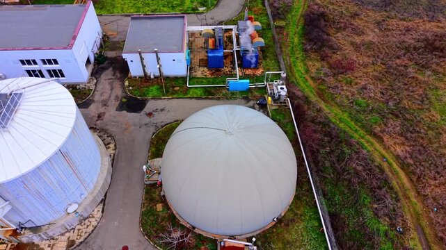 Overhead view of biogas dome and treatment buildings. Aerial perspective of gray inflatable gasholder beside pipework and industrial equipment yard.