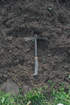 A metal garden hoe rests on freshly turned soil in a garden. The ground is dark and rich, surrounded by green grass.