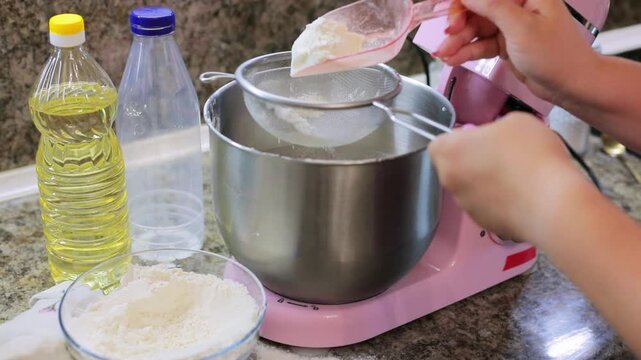 Sifting flour through a sieve into a mixer bowl while preparing homemade dough, an essential baking step in the kitchen alongside cooking oil
