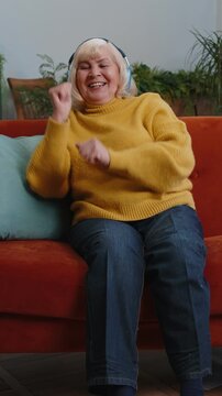 Happy overjoyed senior grandmother woman in wireless headphones relaxing sits on couch at home apartment choosing listening favorite energetic disco dancing music. People weekend leisure activities
