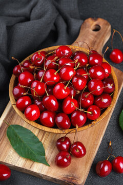 Fresh ripe sweet cherries in wooden bowl on rustic cutting board with green leaves and linen cloth on black textured background. Healthy summer fruit, red berries, fresh seasonal harvest, healthy food
