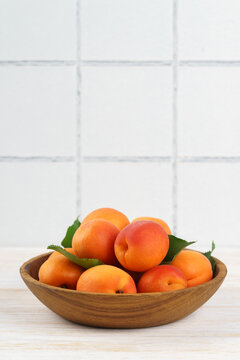 Fresh ripe apricots arranged in a wooden bowl on white kitchen table and tiled wall background with copy space. Summer stone fruits with green leaves, rustic home kitchen setting, healthy food