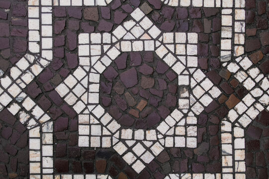 Geometric stone mosaic pavement with contrasting light and dark tiles forming a decorative pattern. Textured cobblestone surface and repeating shapes create an abstract architectural background.