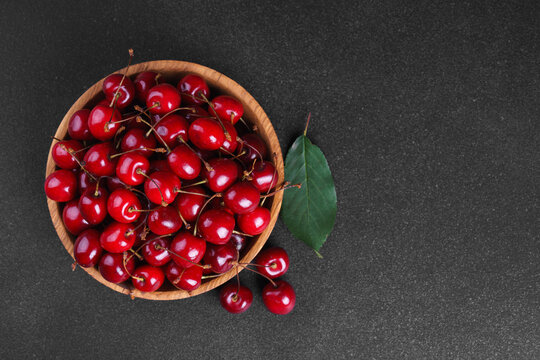 Fresh ripe sweet cherries in rustic wooden bowl with green leaves on black textured background. Top view flat lay with copy space. Juicy organic summer fruit, red berries, healthy seasonal food