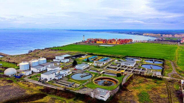 Wide aerial view of coastal wastewater treatment facility, Bulgaria. High angle shows circular clarifiers, storage tanks and dome digester beside beach and resort buildings.