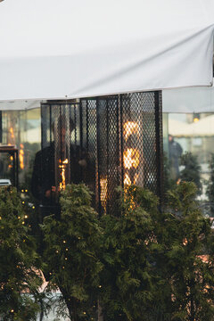Outdoor gas heaters burning behind protective metal grids on a restaurant terrace, decorated with evergreen plants and warm lights. Cozy urban street cafe atmosphere in cold weather.