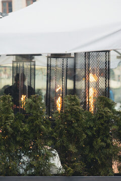 Outdoor gas heaters burning behind protective metal grids on a restaurant terrace, decorated with evergreen plants and warm lights. Cozy urban street cafe atmosphere in cold weather.