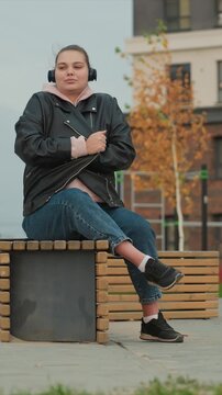 Lady relaxes outdoors clad in casual attire and audio gear. Arms cross under elbow with legs bent to one side off. Setting includes green grass patches distant red plants