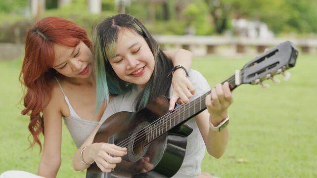 Cheerful Asian lesbian couple playing acoustic guitar and singing together in a sunny green park. Happy young LGBTQ women enjoying romantic musical date and summer freedom.