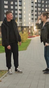 Man in black jacket and pants stands on paved pathway facing. Close up reveals feet of one participant next. Pathway bordered by grass leads to tall residential buildings