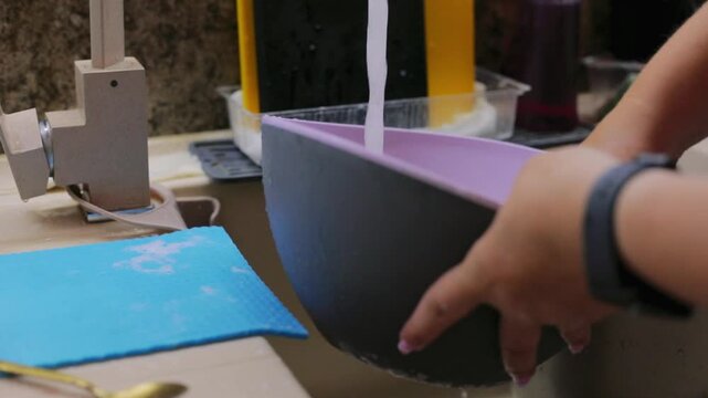 Washing a purple mixing bowl under running water in the kitchen sink with a soapy sponge, cleaning up after homemade baking