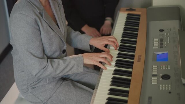 Woman playing piano with teacher, grey suit student focused on keys, coach guiding hand placement, small music school studio, sheet music on stand, calm rehearsal and technique practice