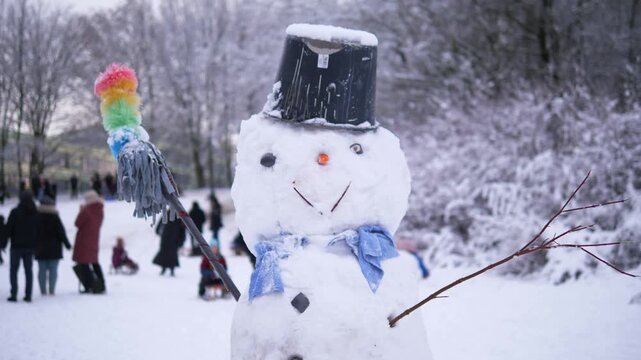 Whimsical snowman with a black hat, blue scarf and colorful broom standing in a snowy winter park with people in the background