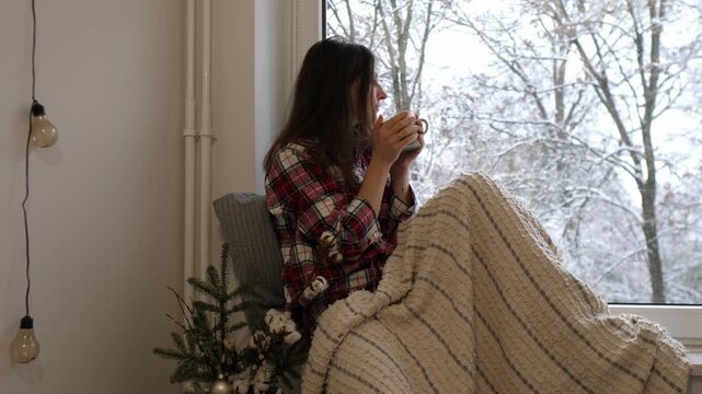 A young woman sitting by the window, wrapped in a warm blanket and holding a cup of hot tea. Snow is falling outside
