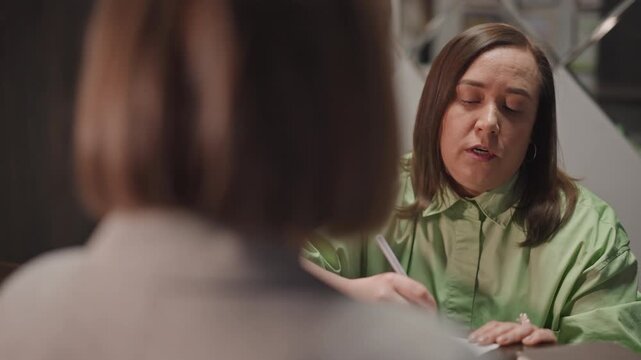 Music teacher reviewing enrollment form in small studio office woman in green jacket takes notes, attentive expression, hands schedule to applicant, warm lighting, calm mentoring tone